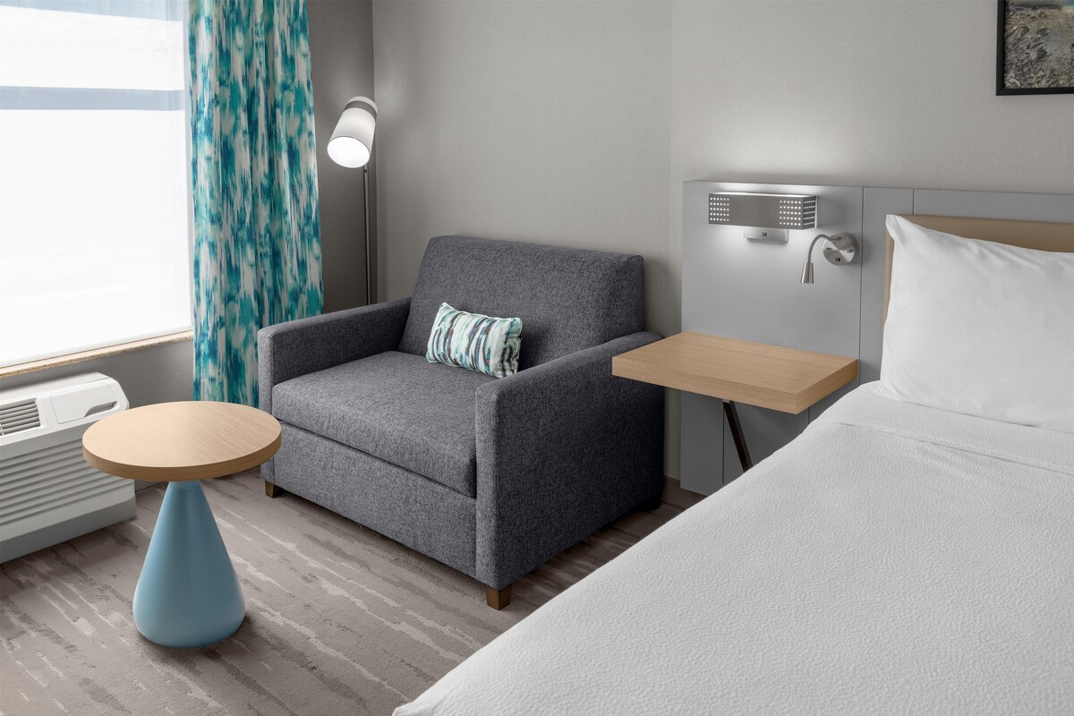 Une chambre d’hôtel avec un fauteuil gris, une petite table ronde, un lit avec une literie blanche, une douche fixée au mur et une fenêtre avec des rideaux.