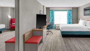 A hotel room with a bed, a desk with a TV, a chair, a bench, and a window with curtains.