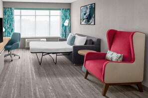 Una habitación de hotel con una cama, un sillón rojo, un sofá gris, un sillón azul, una mesita redonda pequeña, un escritorio, una lámpara y una ventana con cortinas.