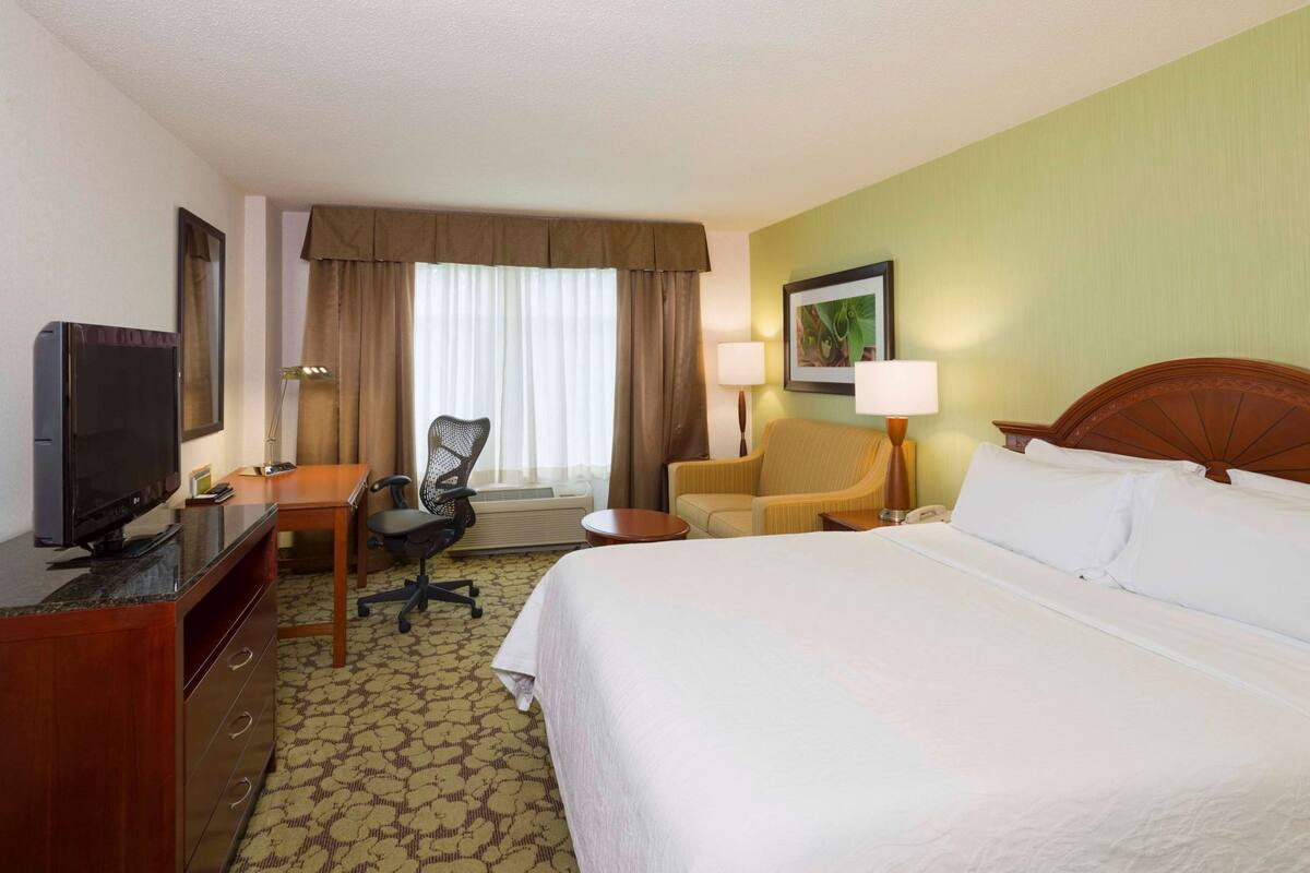 A hotel room with a bed, a desk with a TV, a chair, a lamp, and a window with curtains.