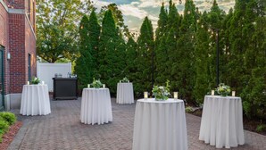 A series of round tables with white tablecloths, arranged outdoors on a brick-paved area with a backdrop of trees and a building. - Hilton Garden Inn Hartford South/Glastonbury (Glastonbury)
