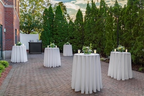 A series of round tables with white tablecloths, arranged outdoors on a brick-paved area with a backdrop of trees and a building. - Hilton Garden Inn Hartford South/Glastonbury (Glastonbury)