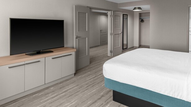 A hotel room with a bed, a TV on a wooden console, a door leading to a bathroom, and a hallway.