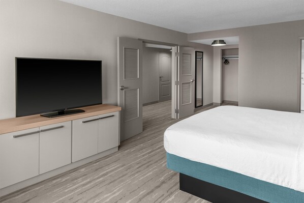 Una habitación de hotel con cama, un televisor sobre una consola de madera, una puerta que da al baño y un pasillo.