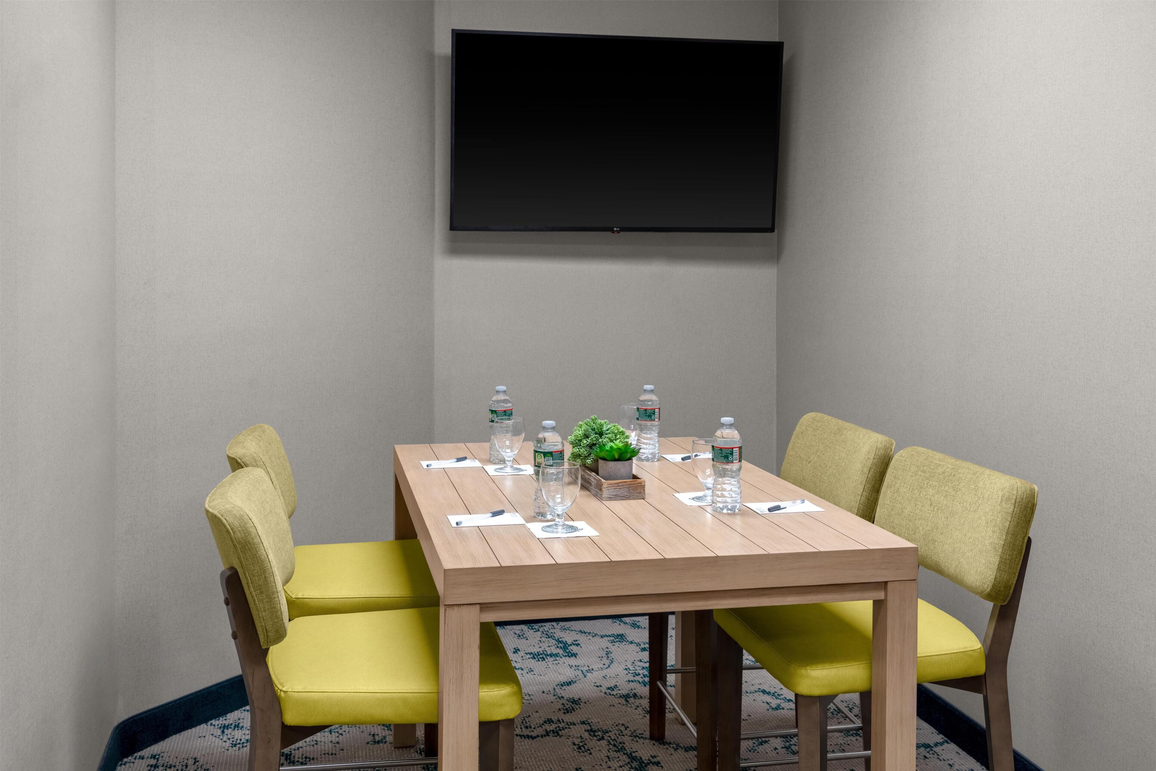 Un salón de conferencias con una mesa de madera, sillas amarillas, un televisor de pantalla plana en la pared y botellas de agua sobre la mesa.