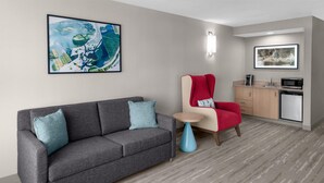 A modern hotel room with a gray sofa, a red chair, a small round table, a kitchenette, and two framed artworks on the walls. - Hilton Garden Inn Hartford South/Glastonbury (Glastonbury)