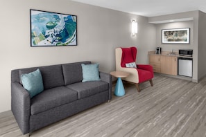 A modern hotel room with a gray sofa, a red chair, a small round table, a kitchenette, and two framed artworks on the walls. - Hilton Garden Inn Hartford South/Glastonbury (Glastonbury)