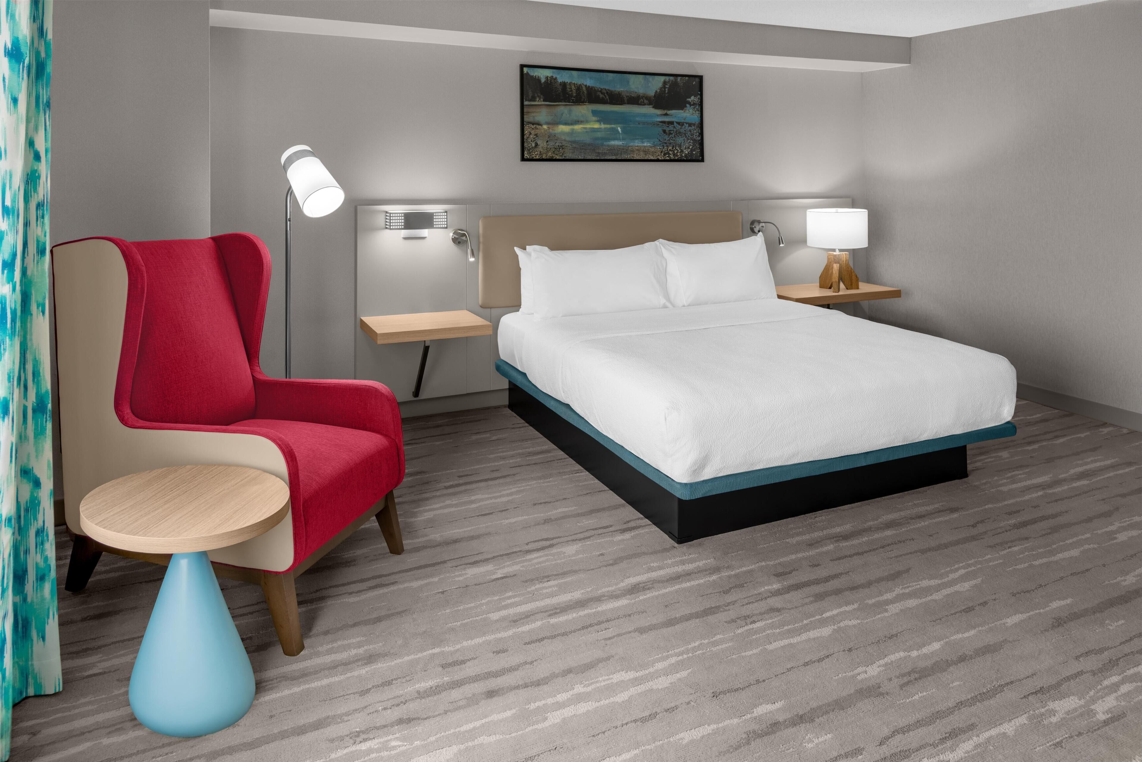 A hotel room with a bed, a red armchair, a small round table, two bedside lamps, and a painting on the wall.
