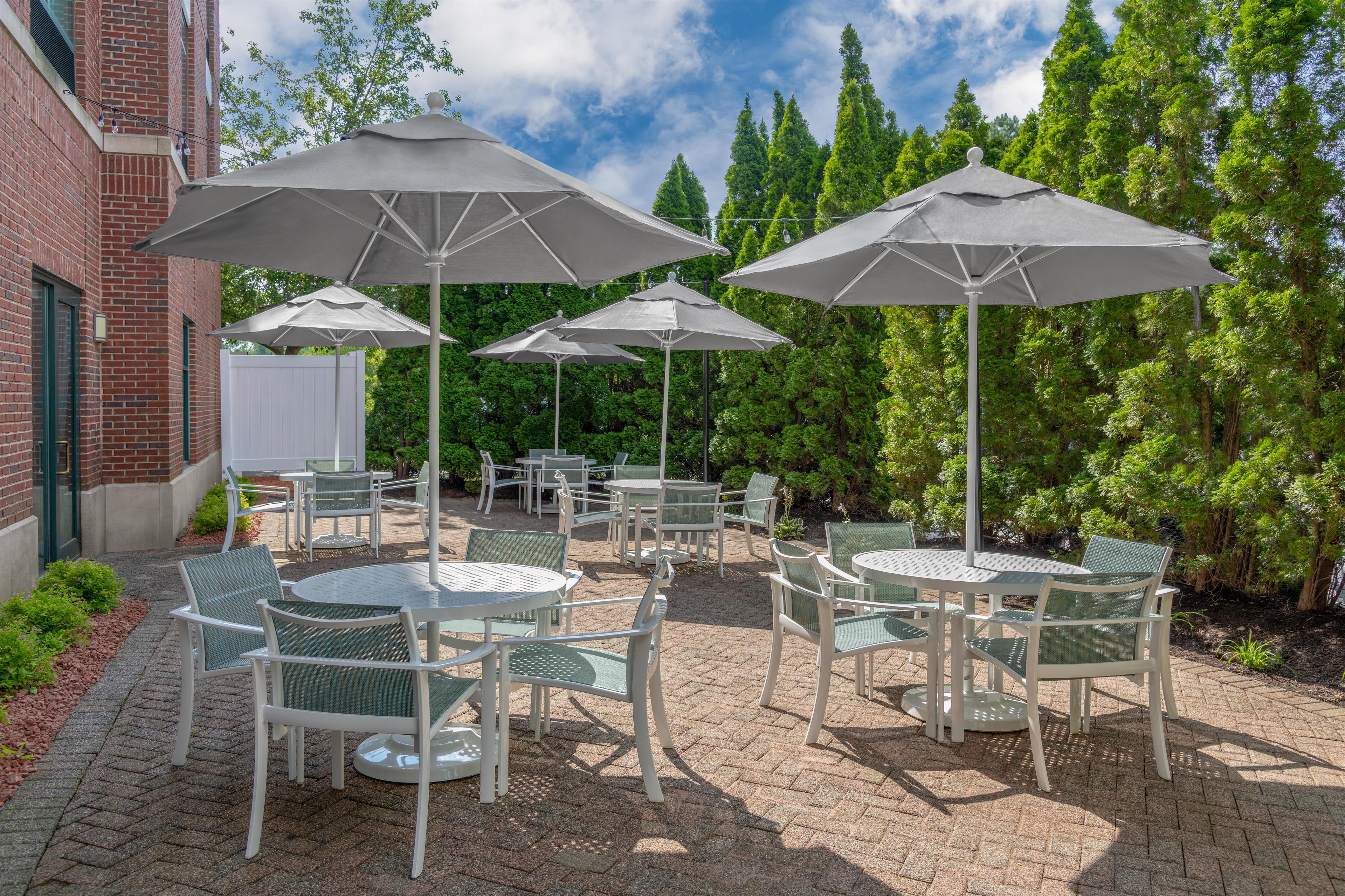 An outdoor patio with round tables and chairs, shaded by large umbrellas, set against a backdrop of trees and a brick building.