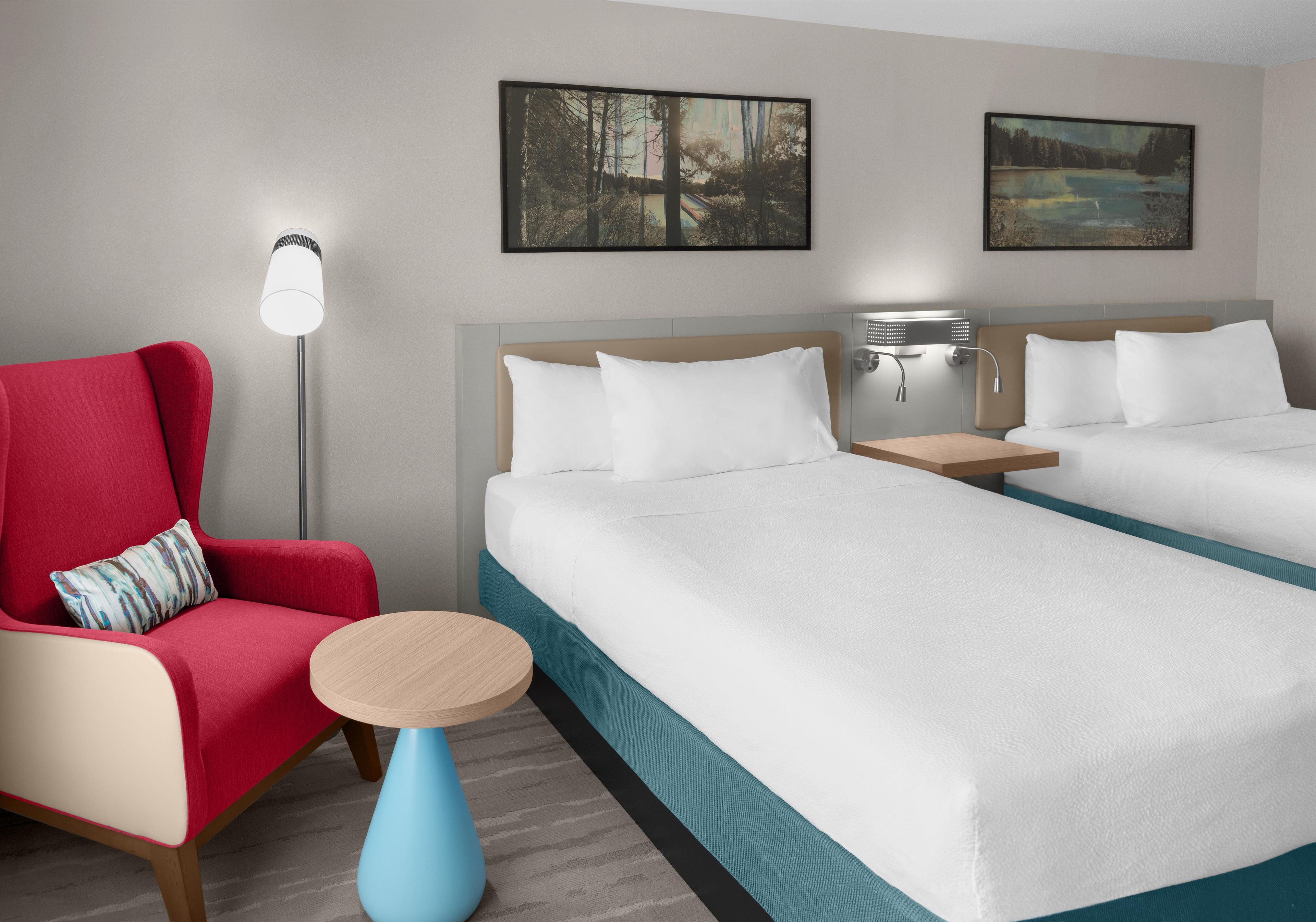 A hotel room with two beds, a red armchair, a small round table, and two wall-mounted lamps.