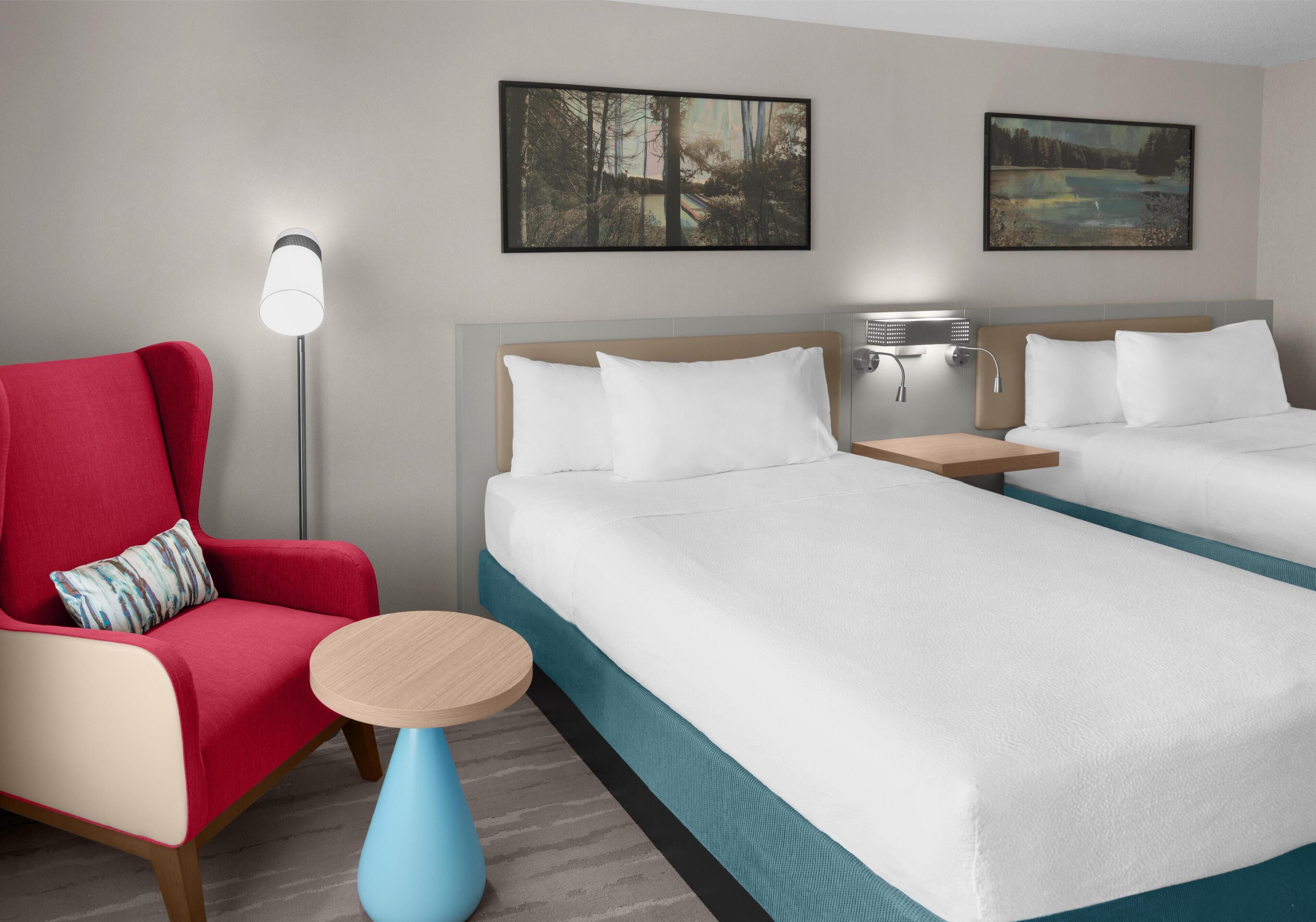 A hotel room with two beds, a red armchair, a small round table, and two wall-mounted lamps.