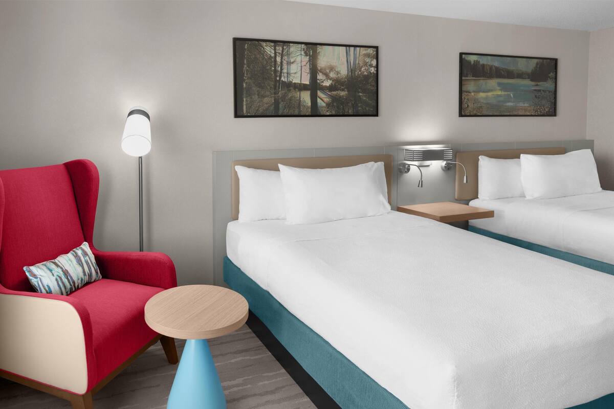 Habitación de hotel con dos camas, un sillón rojo, una mesita redonda y dos lámparas fijadas a la pared.