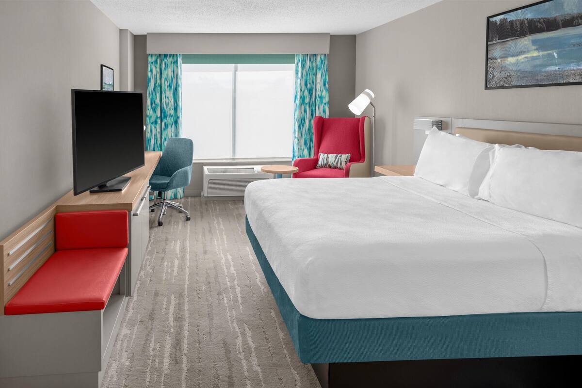 Habitación de hotel con cama, escritorio con televisor, silla, lámpara de escritorio y ventana con cortinas.