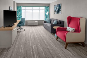 Habitación de hotel con televisor de pantalla plana, escritorio, silla de oficina azul, sofá gris, sillón rojo y ventana grande con cortinas.