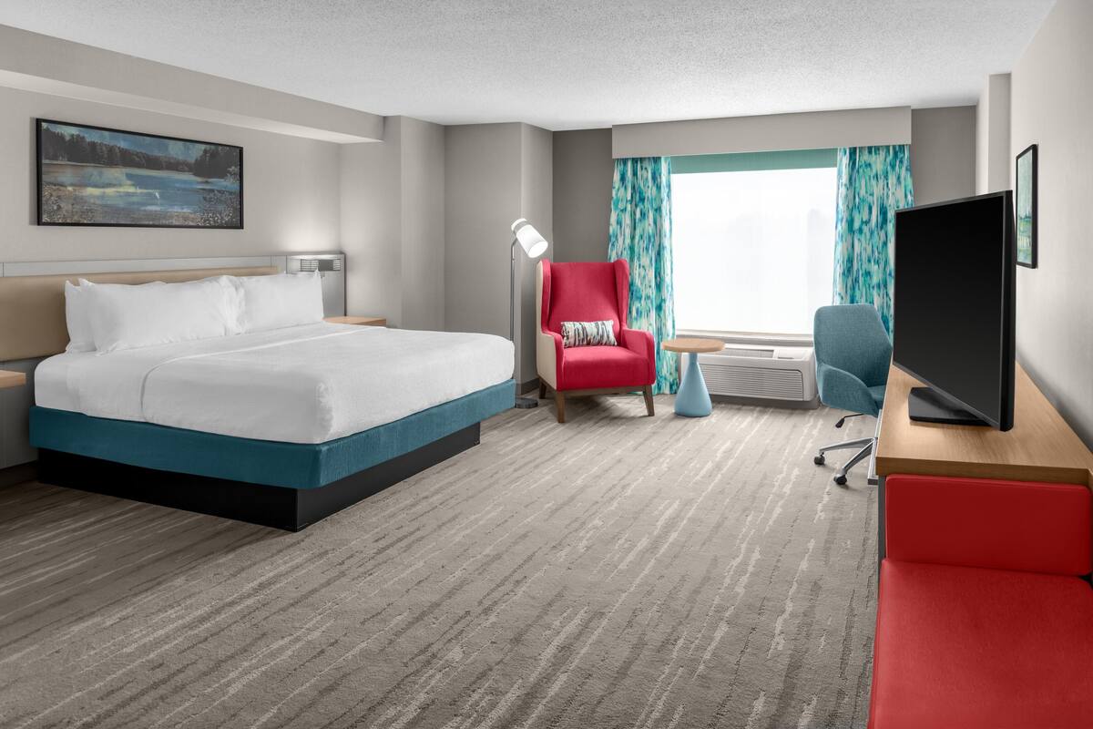 Una habitación de hotel con cama, un sillón rojo, televisor y ventana con cortinas.