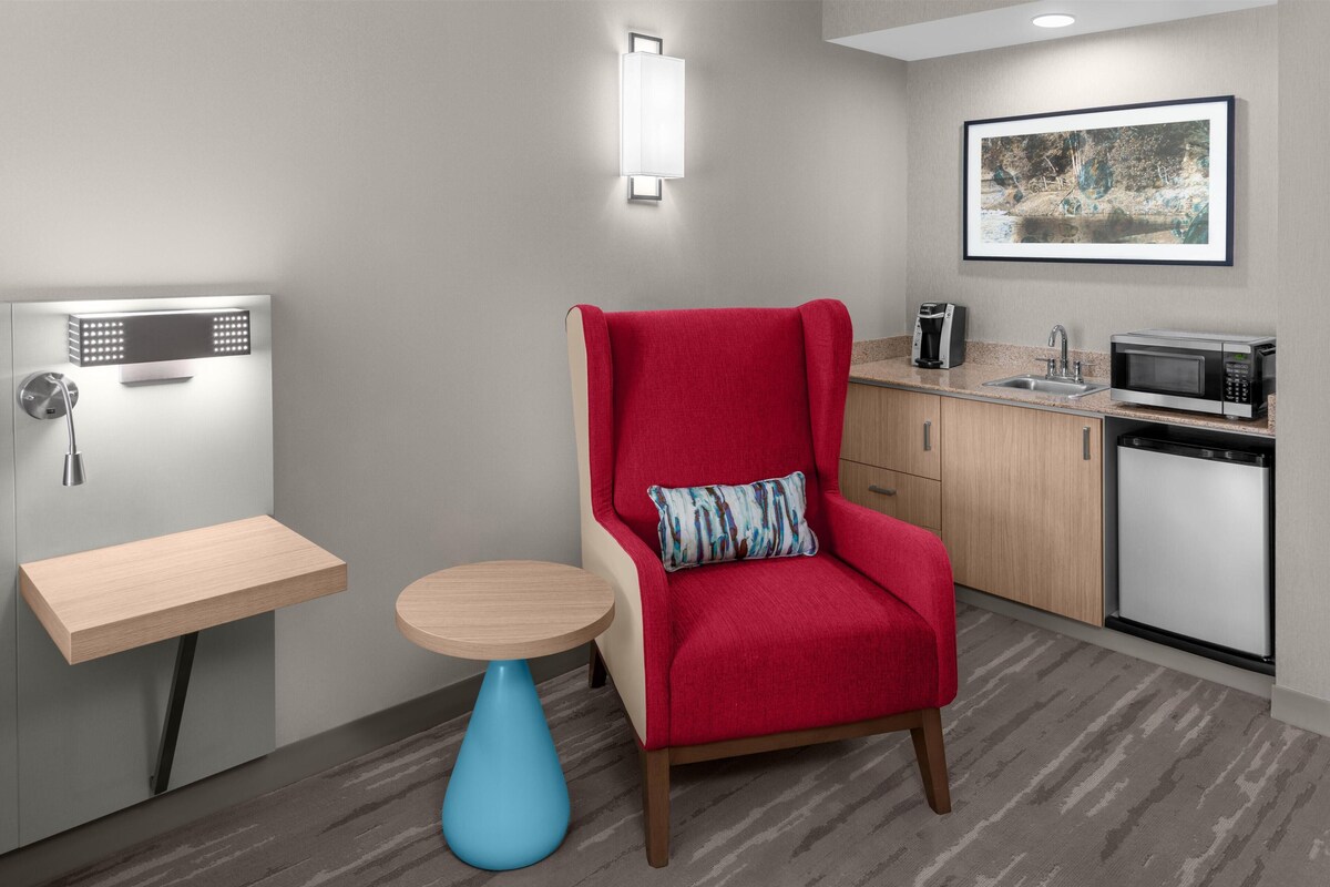 Habitación de hotel moderna con un sillón rojo, una mesita redonda pequeña, microondas, lavamanos y un cuadro enmarcado en la pared.