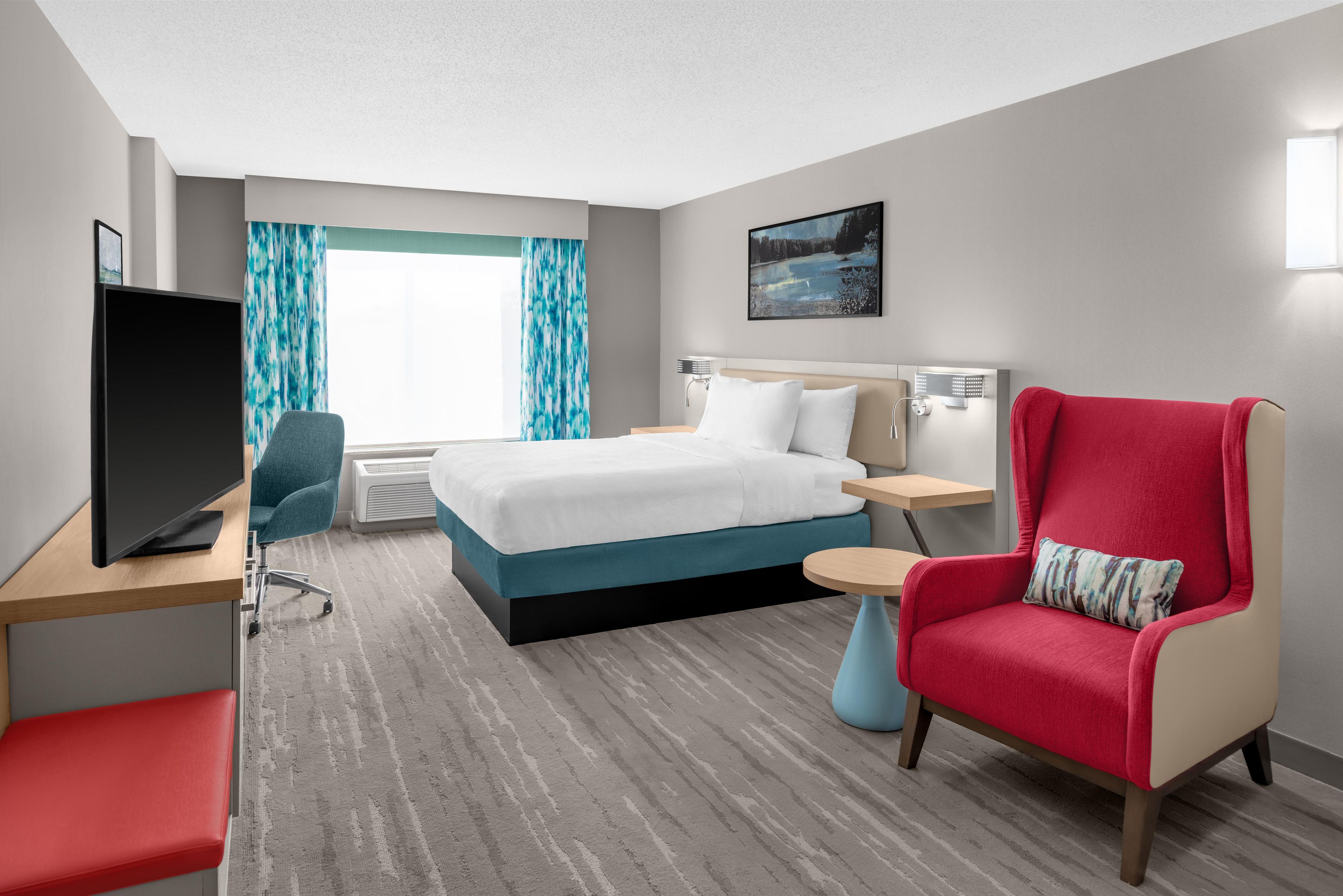 A hotel room with a bed, a TV, a desk, a chair, a small table, and a window with curtains.
