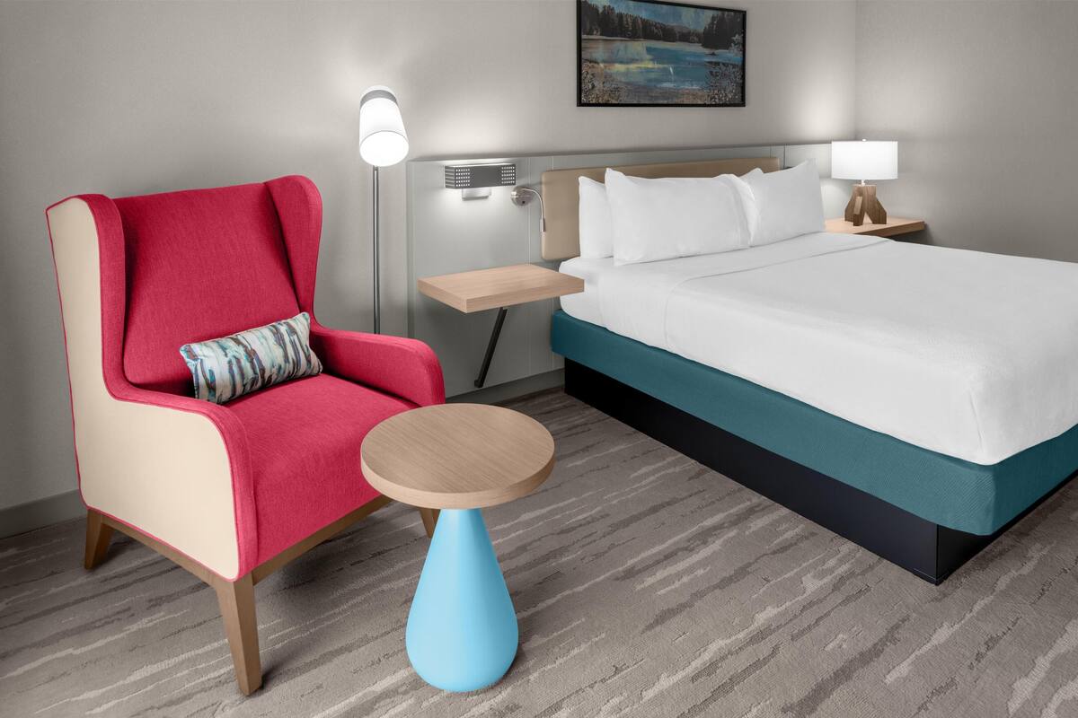 Una habitación de hotel con una cama, un sillón rojo, una mesita azul, una mesita de noche con una lámpara y una luz empotrada en la pared.