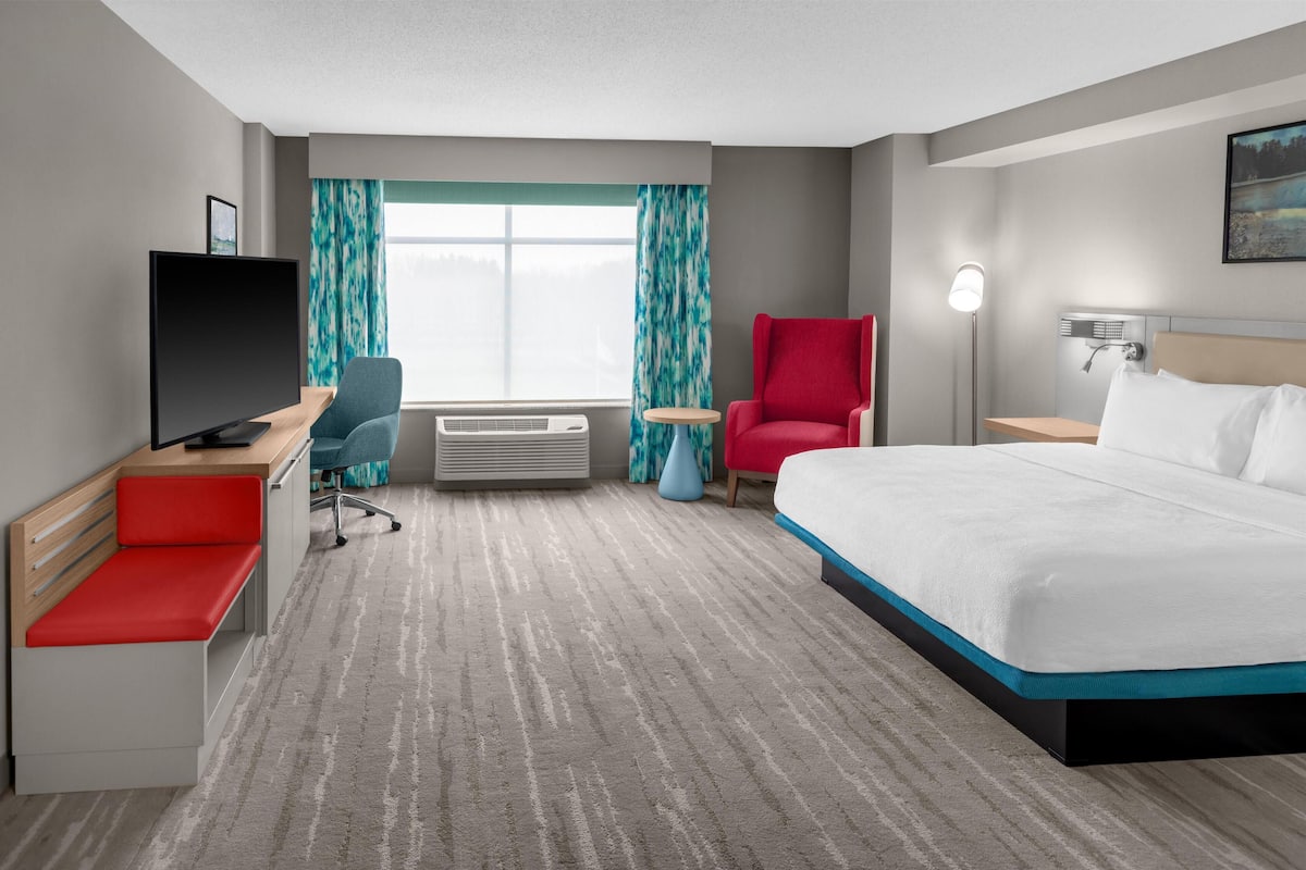 Una habitación de hotel con cama, escritorio con televisor, silla roja, mesita y ventana con cortinas.