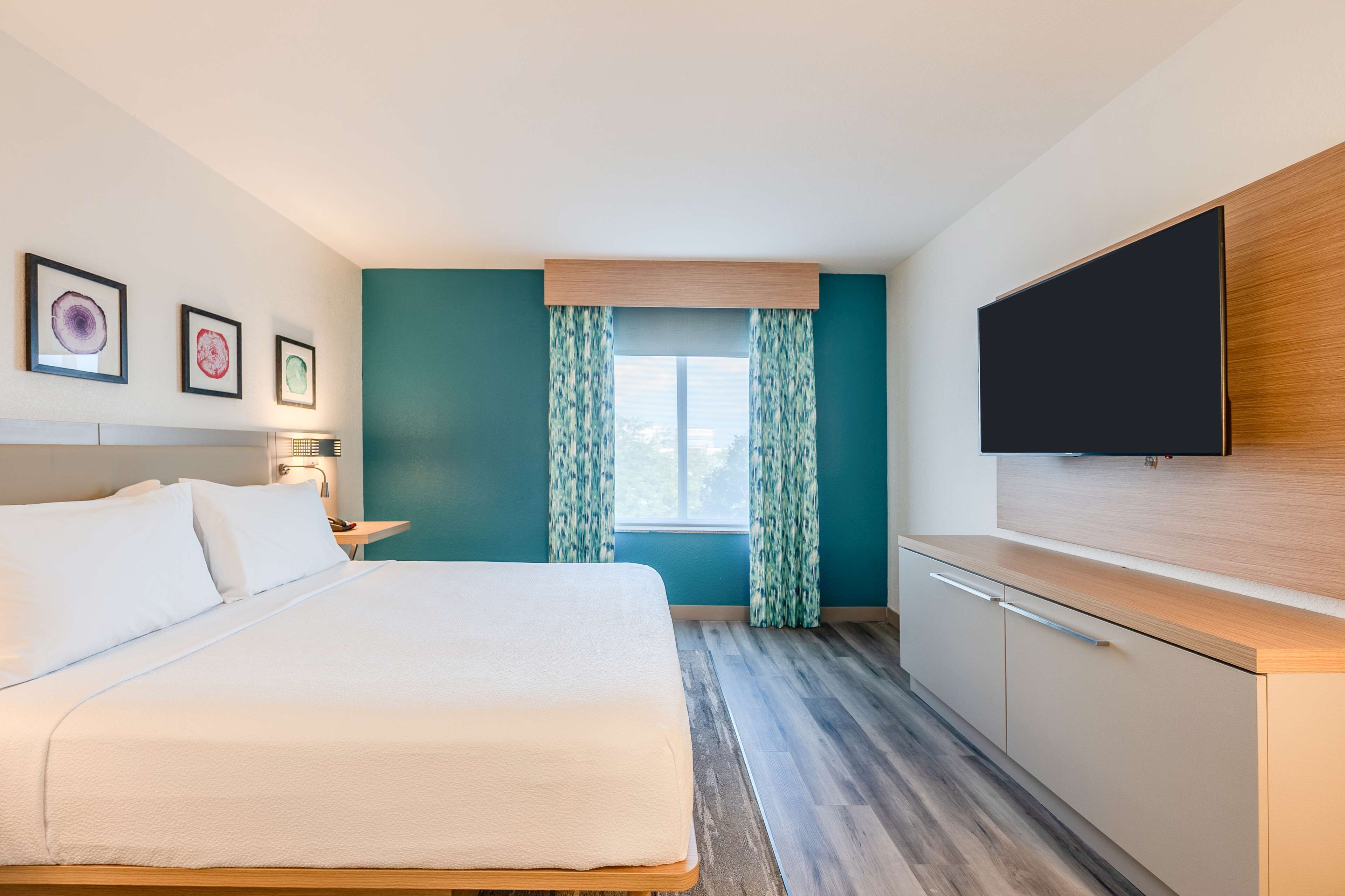 Une chambre d’hôtel avec un lit, un téléviseur fixé au mur, une fenêtre avec des rideaux et des œuvres d’art accrochées au mur.