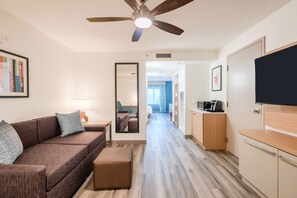 A modern hotel room with a brown sofa, a small table with a lamp, a ceiling fan, and a TV on a wooden cabinet. - Hilton Garden Inn Fort Lauderdale Airport-Cruise Port (Dania Beach)