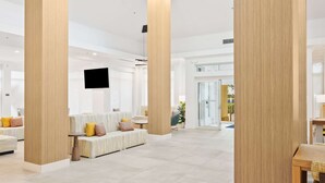 Un amplio vestíbulo de hotel con techos altos, un área de estar central con sofás y sillones, y un televisor de pantalla plana grande montado en la pared.