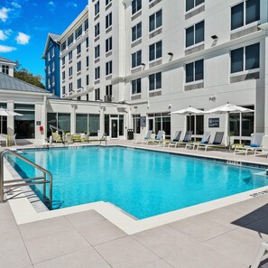 A swimming pool with lounge chairs, a lifeguard sign, and a building with multiple windows and balconies. - Hilton Garden Inn Fort Lauderdale Airport-Cruise Port (Dania Beach)