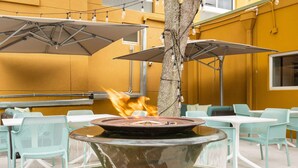 Un área de comedor al aire libre con una chimenea central, rodeada de mesas y sillas, y sombrillas que proporcionan sombra.