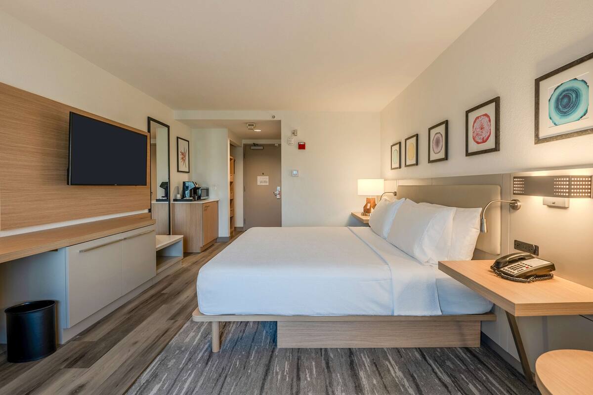 A hotel room with a bed, bedside table, lamp, and a flat-screen TV mounted on the wall.