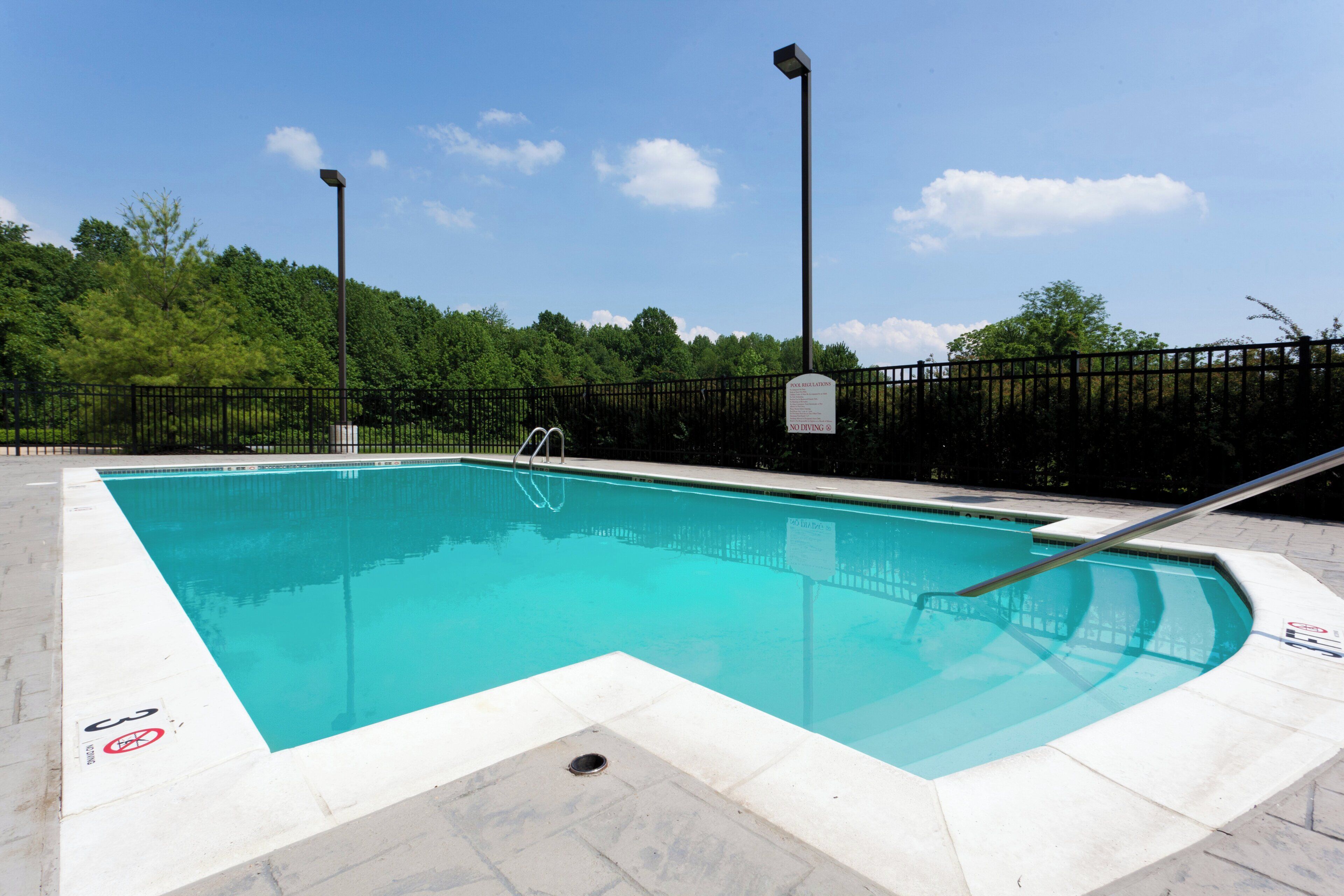 A rectangular swimming pool with a metal ladder and a no diving sign, surrounded by a black fence and trees in the background.