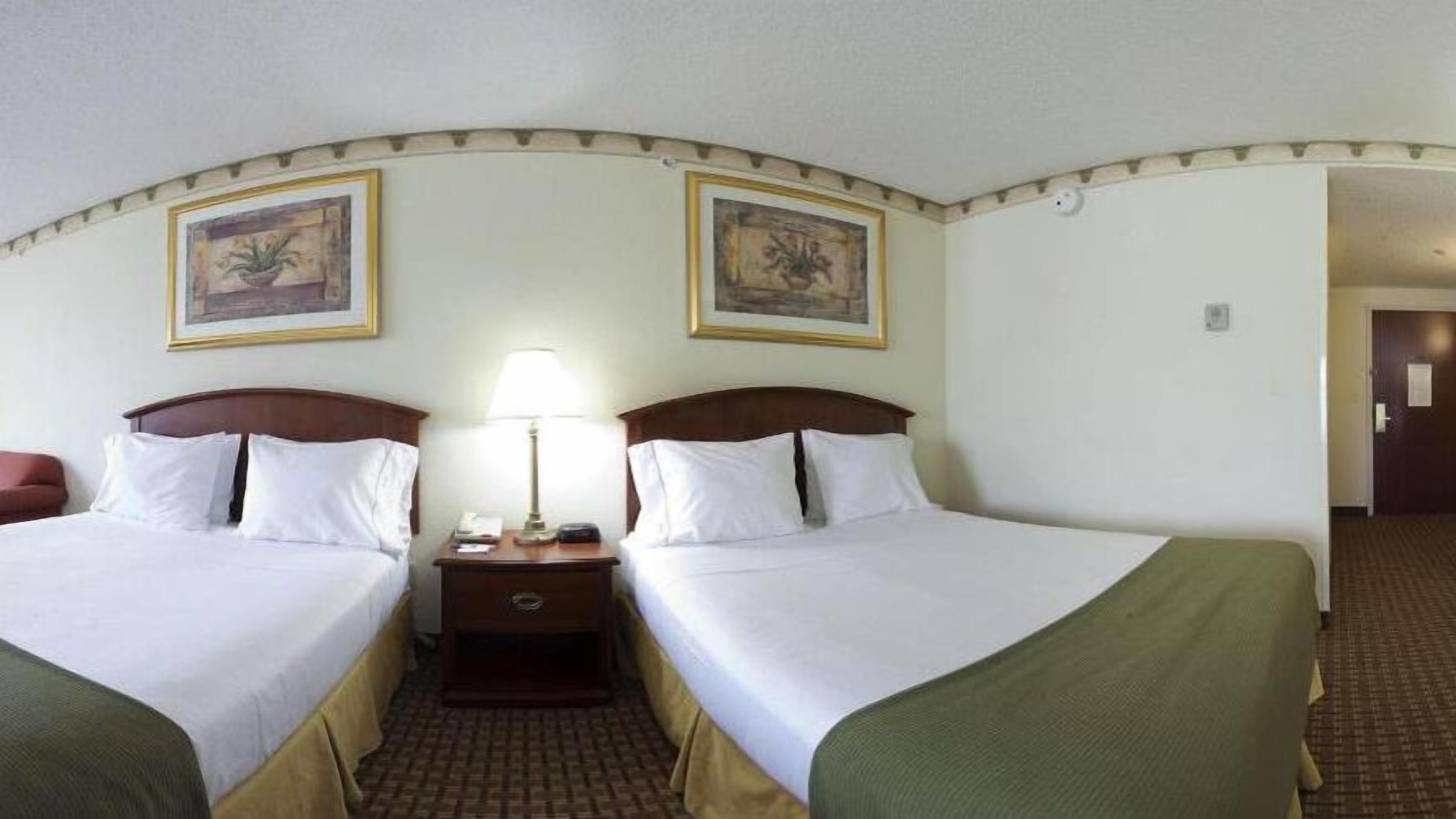Habitación de hotel con dos camas, una mesita de noche con lámpara y dos cuadros en la pared.