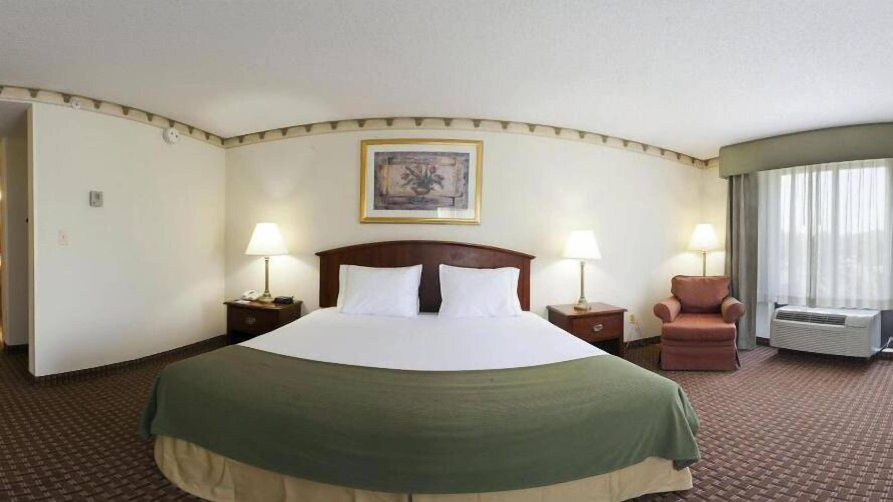 A hotel room with a large bed, two bedside tables with lamps, a chair, a window with curtains, and a painting on the wall.