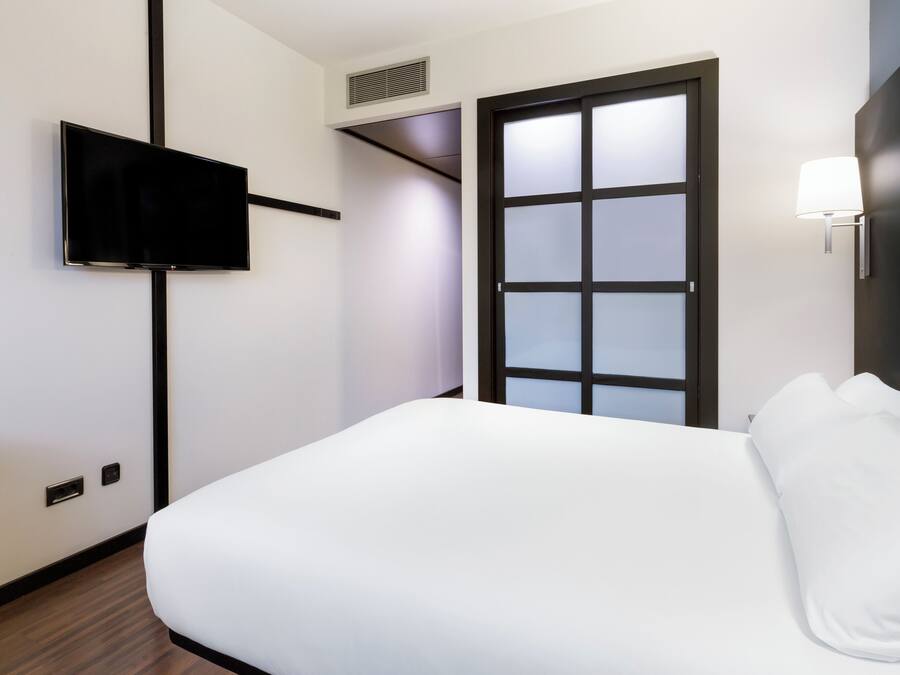 Habitación de hotel con cama, televisión de pantalla plana en la pared, ventana con vidrio esmerilado y lámpara fijada a la pared.