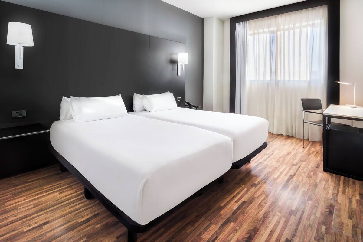 A hotel room with two beds, a desk, a chair, and a window with curtains.