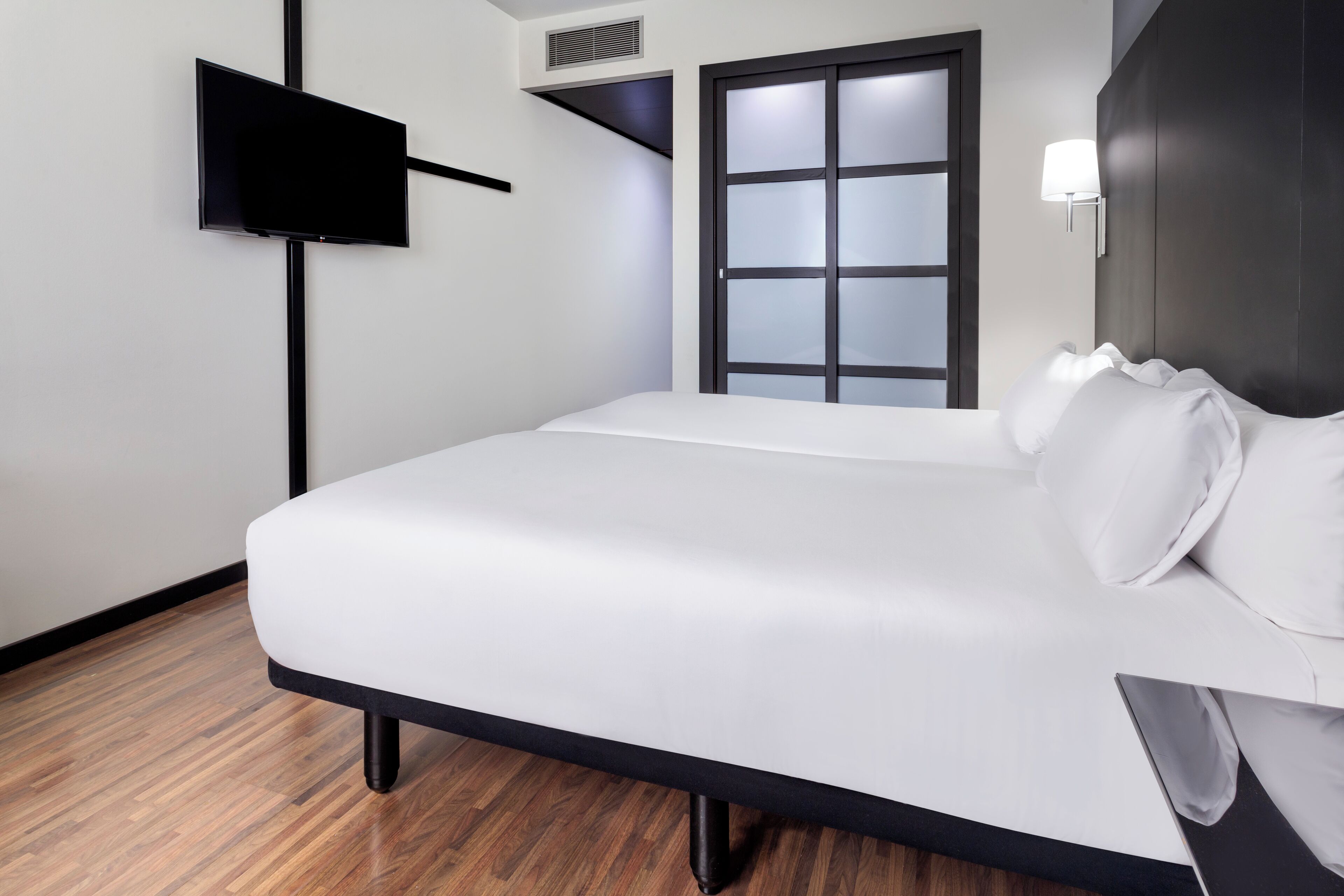 A modern hotel room with a large bed, a flat-screen TV mounted on the wall, a window with frosted glass, and a wall-mounted lamp.