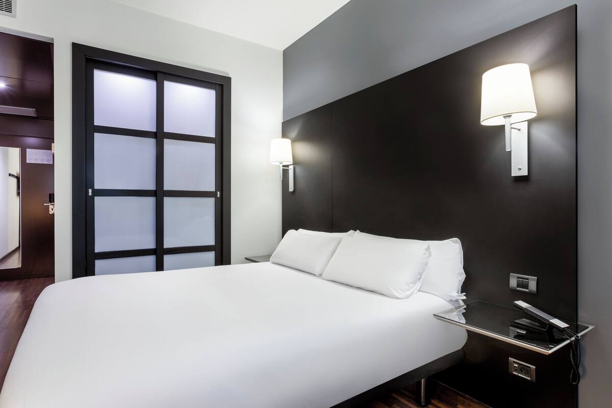 A modern hotel room with a large bed, a dark headboard, a wall-mounted lamp, and a bedside table with a phone.