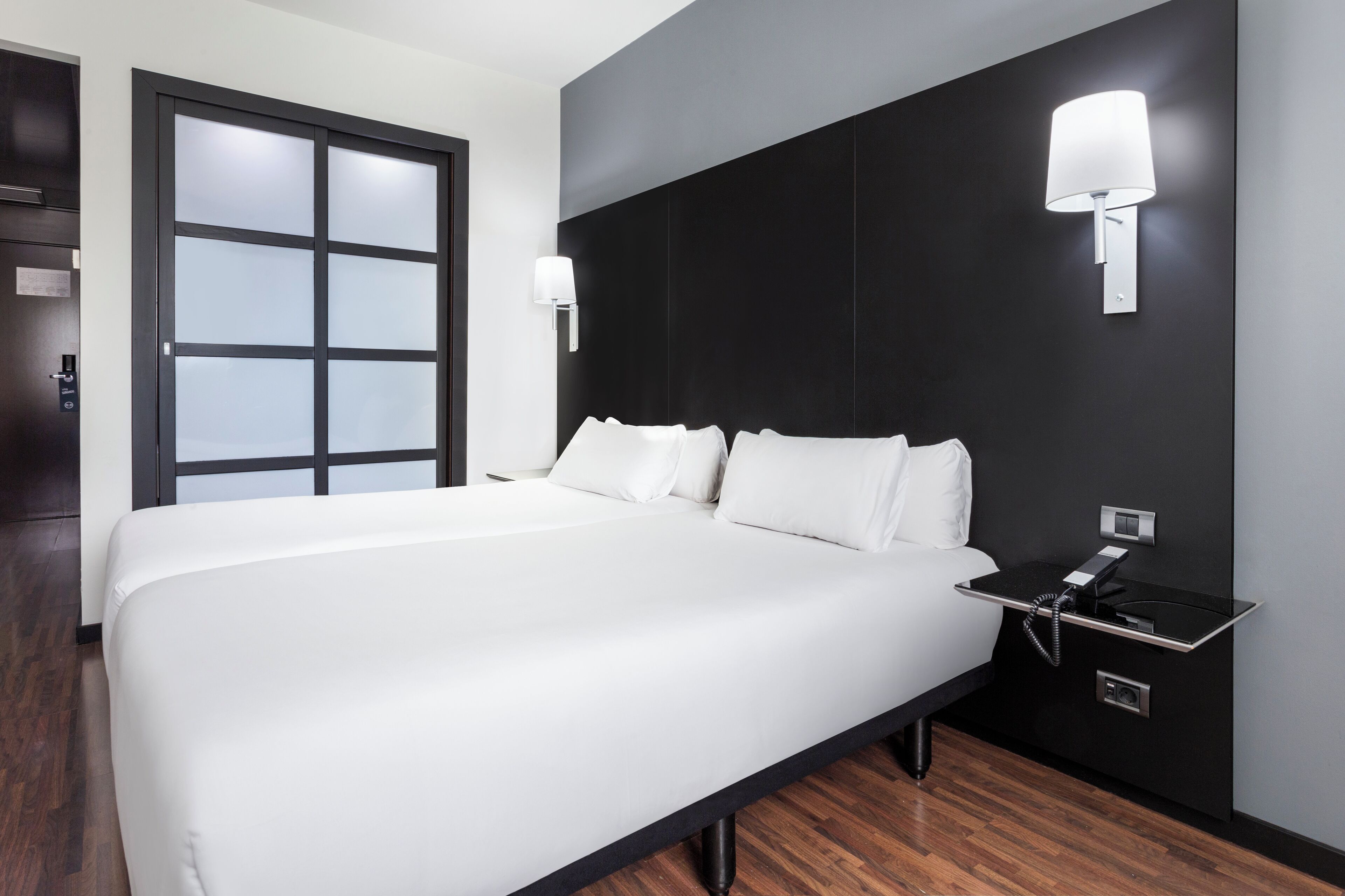 A modern hotel room with a large bed, a bedside table with a phone, and a wall-mounted lamp.