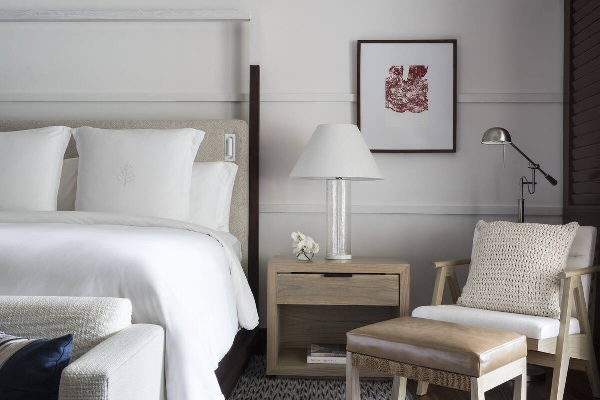 A neatly arranged hotel room with a bed, a nightstand with a lamp, a chair, and a framed artwork on the wall.
