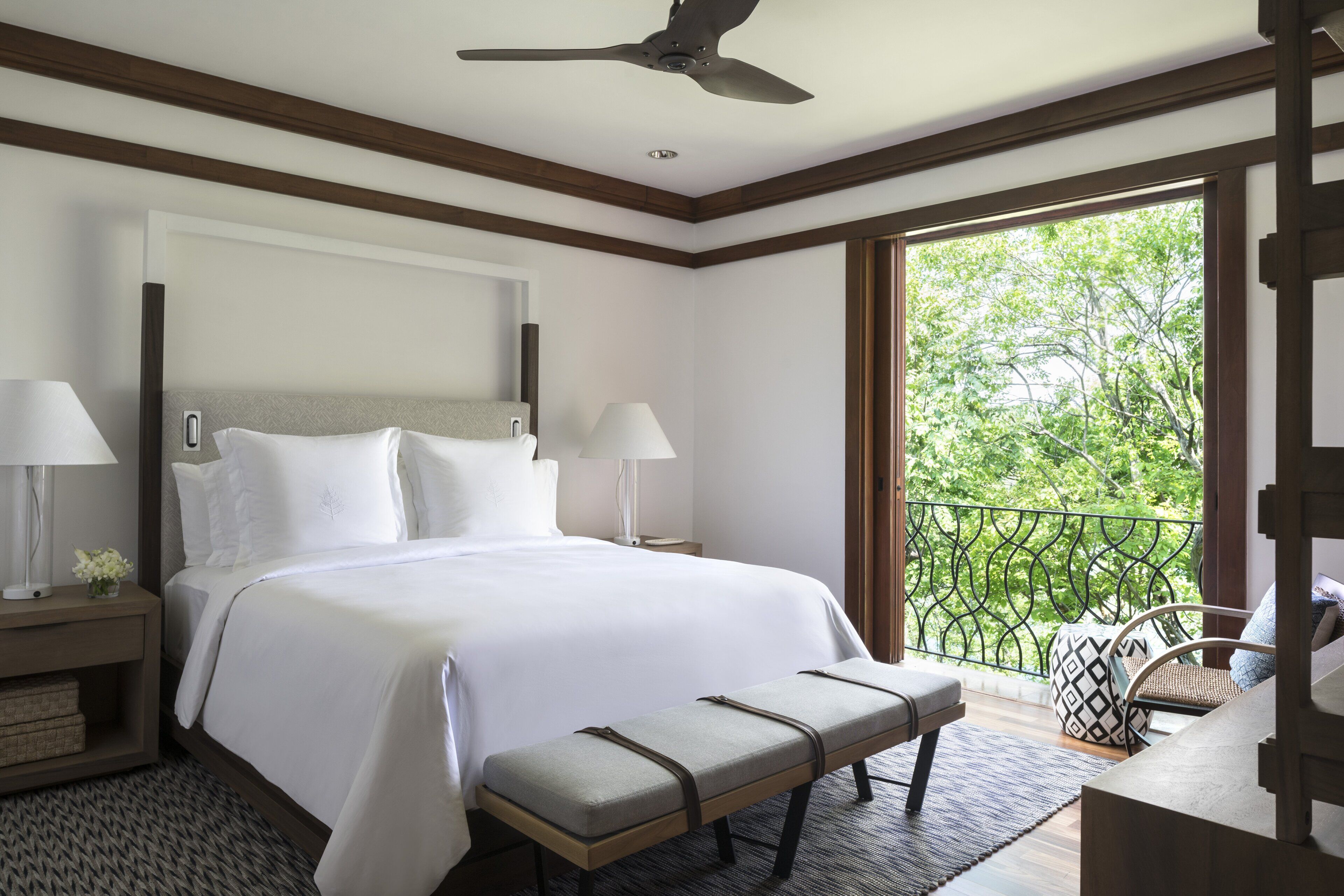 A hotel room with a large bed, two bedside tables with lamps, a bench, and a balcony with a view of greenery.