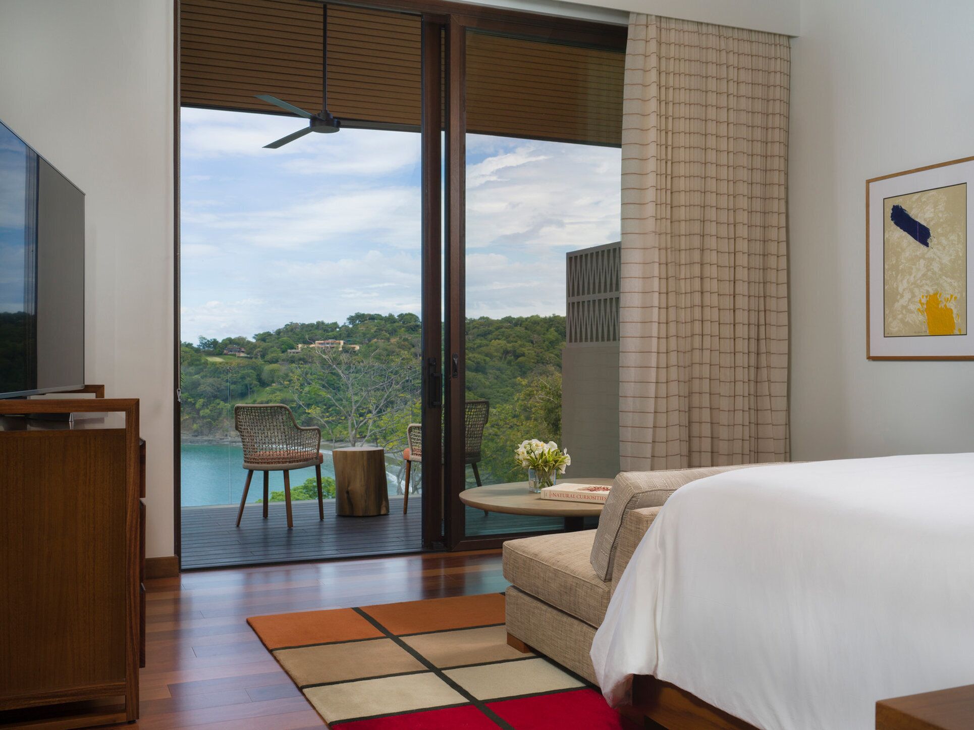 A bedroom with a large bed, a TV, and a view of a lush landscape through a sliding glass door.