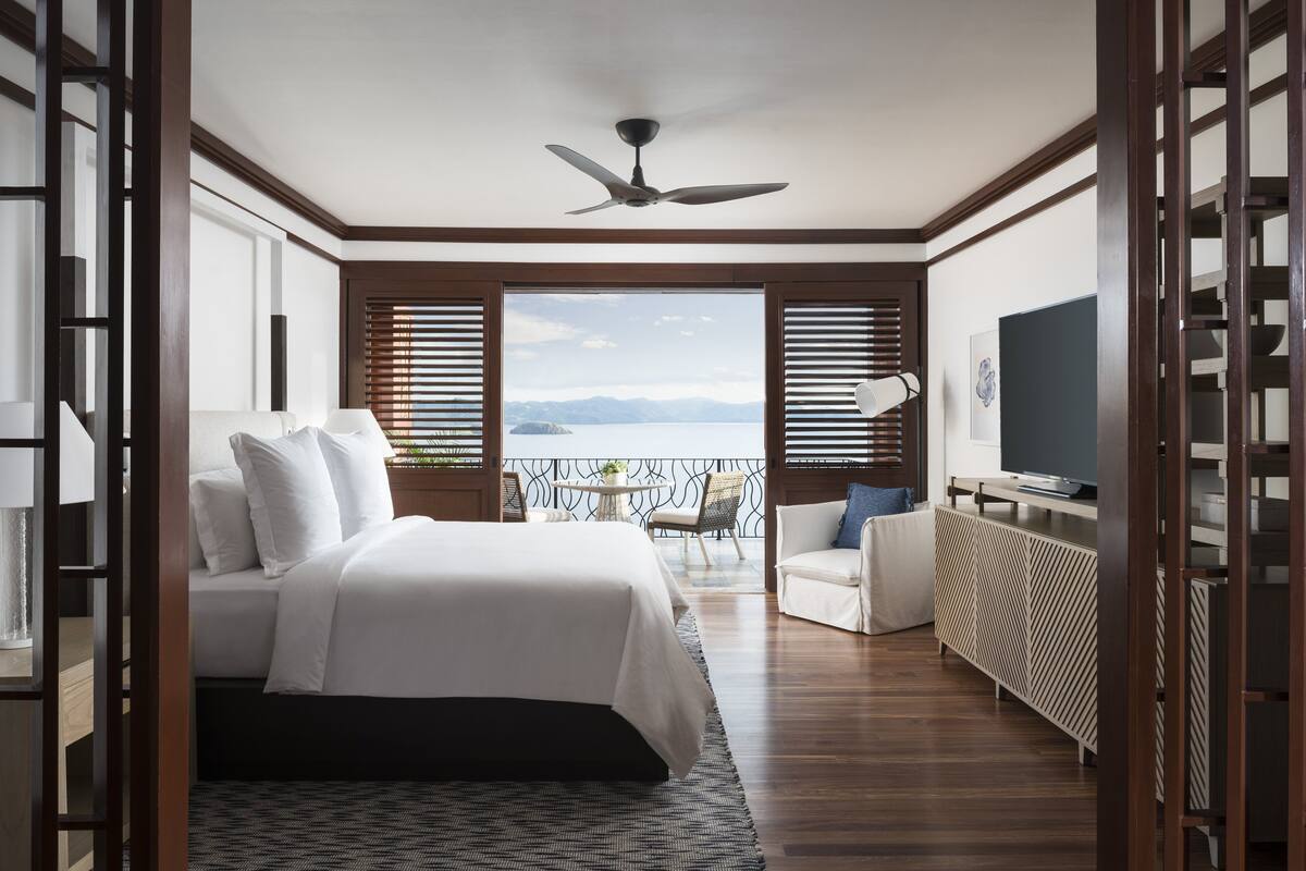 A bedroom with a bed, a desk, a chair, a TV, and a balcony with a view of the sea.