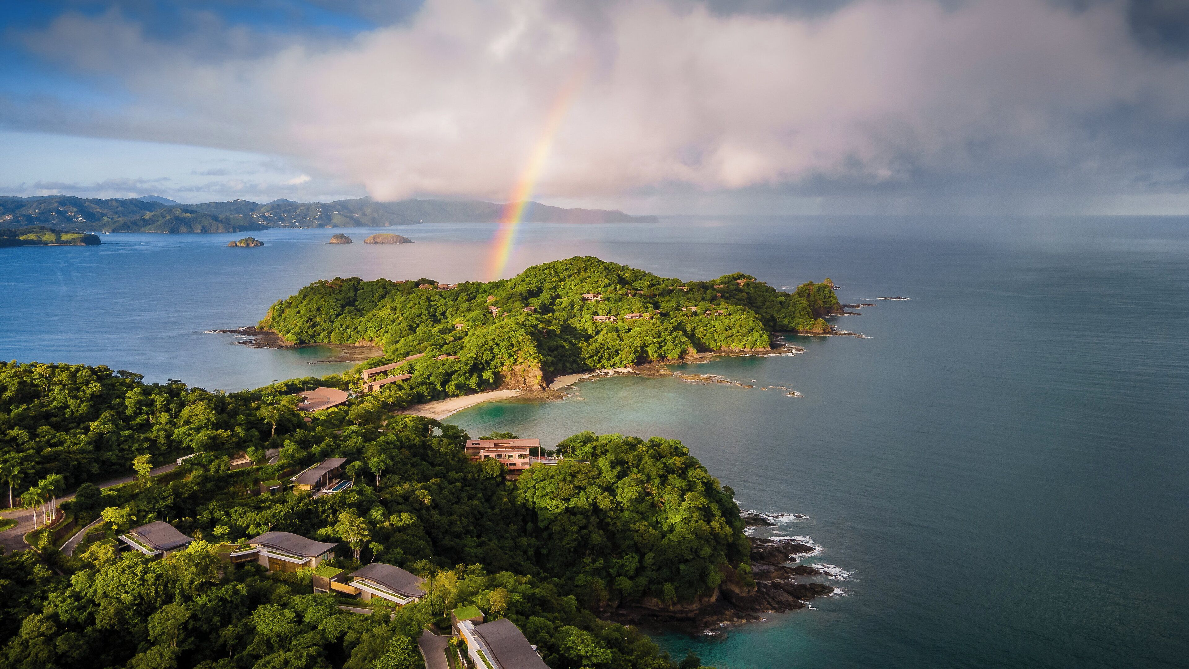 An aerial view of a tropical island with lush greenery, clear blue waters, and a visible rainbow.