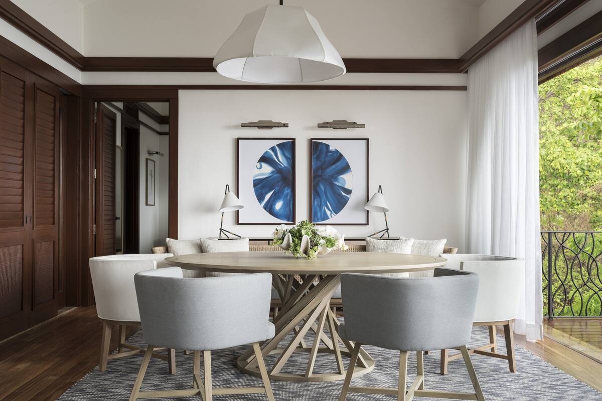A dining area with a round table, six chairs, two wall-mounted lamps, and two framed blue floral artworks.