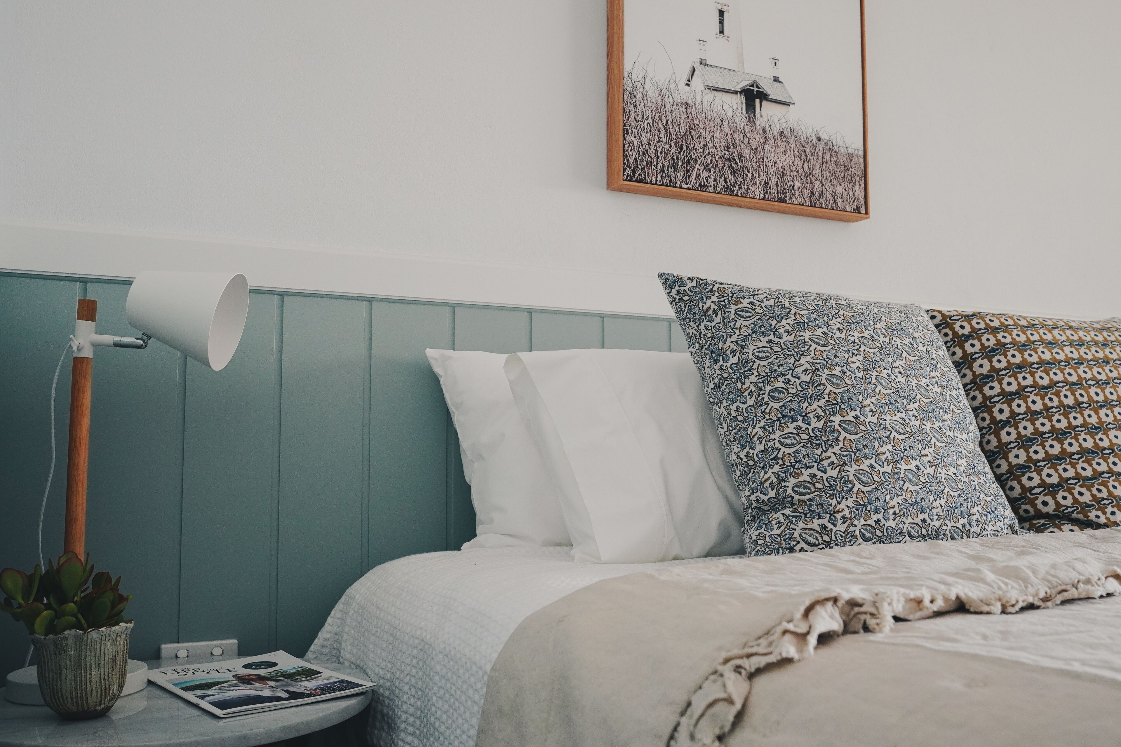 Une chambre à coucher bien rangée, avec un lit, des oreillers, une lampe de chevet, une petite table sur laquelle est posée une plante, et un tableau encadré accroché au mur.
