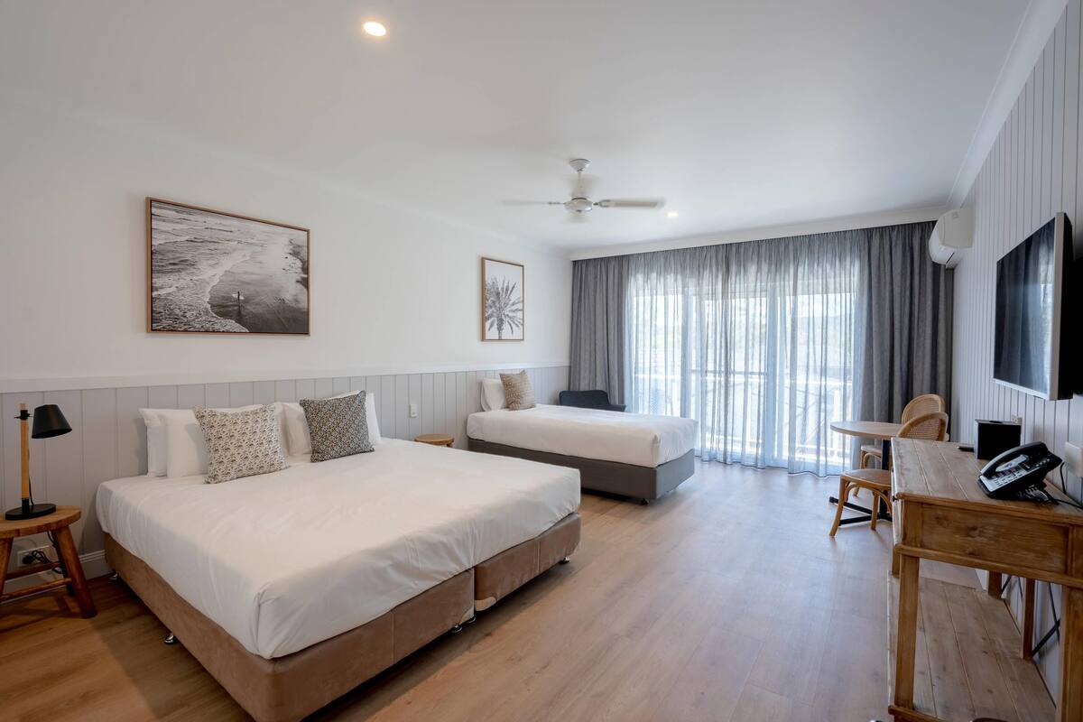 A hotel room with a large bed, a smaller bed, a desk with a phone, a chair, a TV, and a ceiling fan.