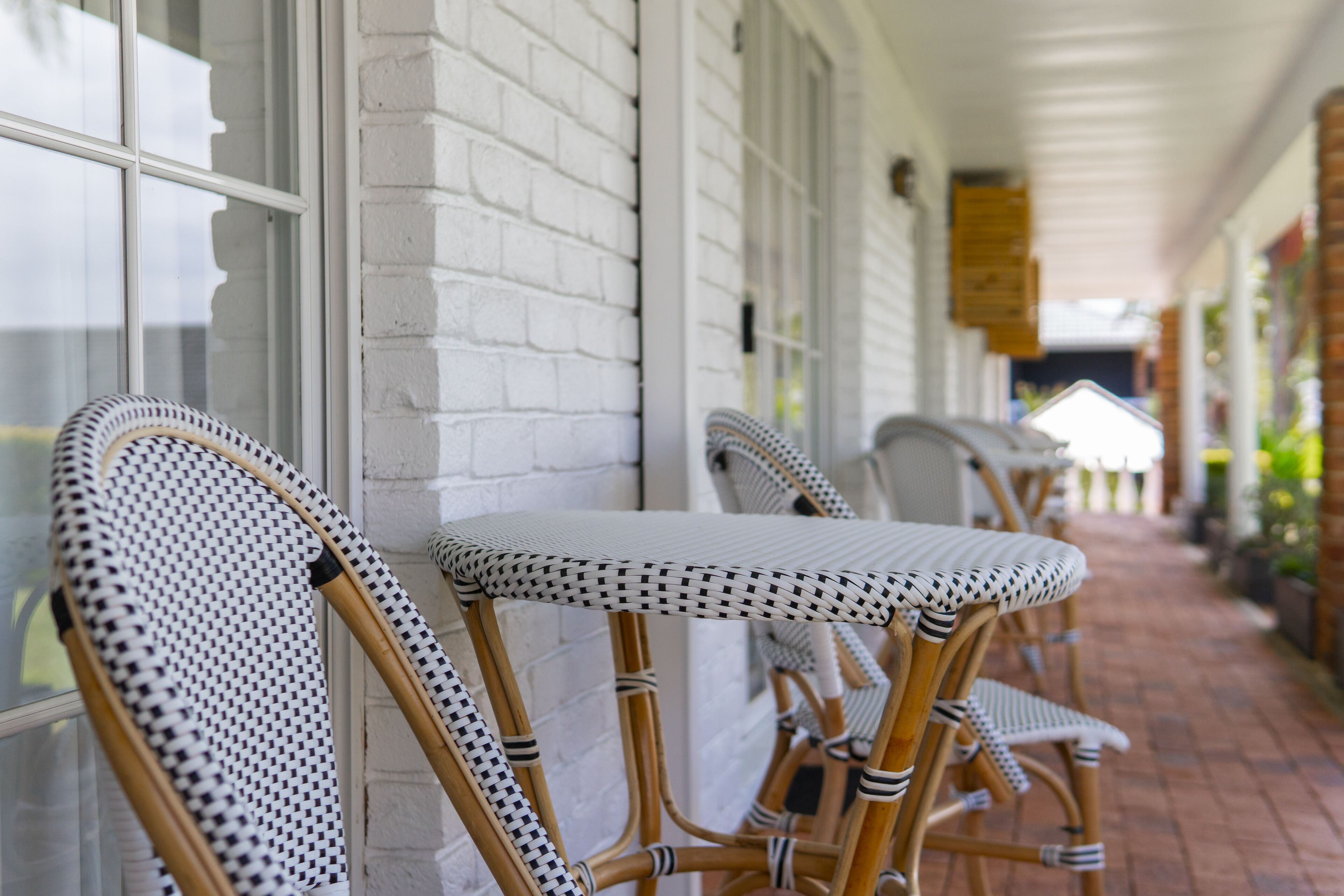 Wicker chairs with checkered cushions aligned against a white brick wall on a brick-paved walkway.