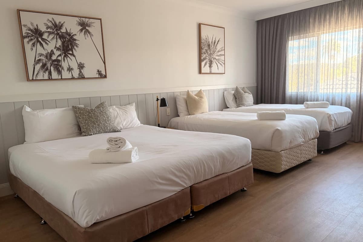 A hotel room with three beds, a wooden floor, and two framed pictures on the wall.