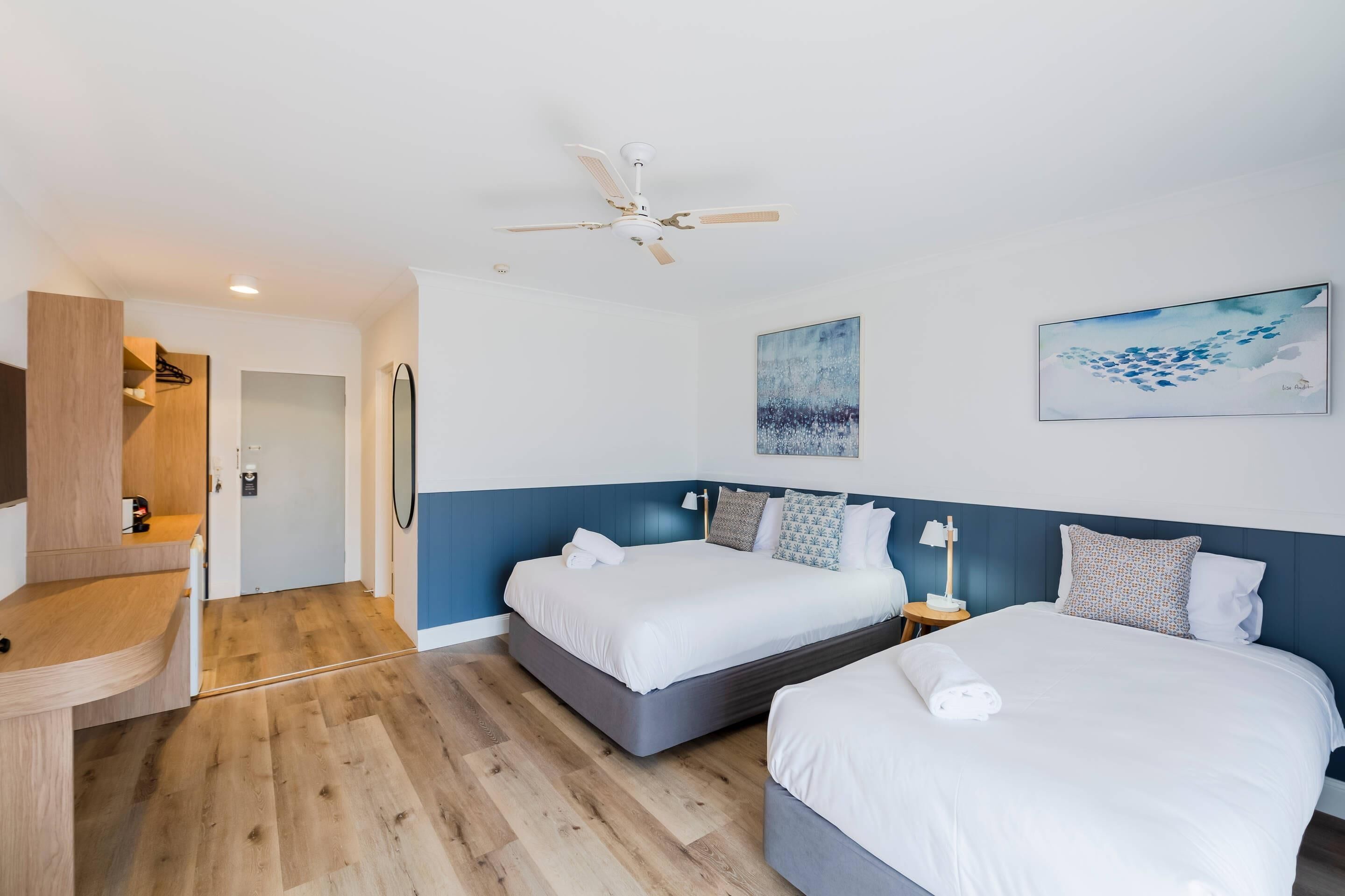 A hotel room with two beds, a ceiling fan, artwork on the walls, and a wooden desk.