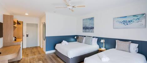 A hotel room with two beds, a ceiling fan, artwork on the walls, and a wooden desk.