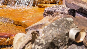Un arroyo pedregoso con agua corriendo sobre las rocas, una cerca blanca y un árbol al fondo.